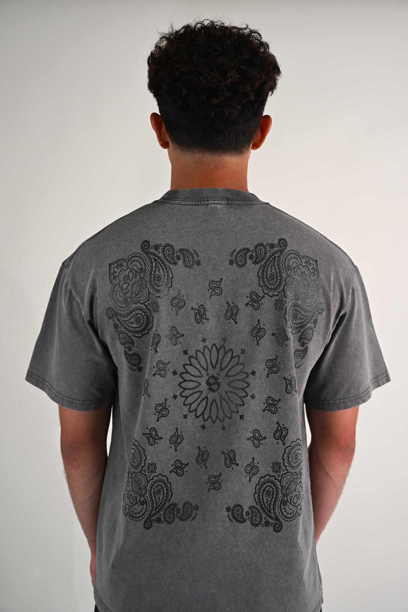 Person wearing a gray t-shirt with black paisley calavera bandana design on the back against a plain background