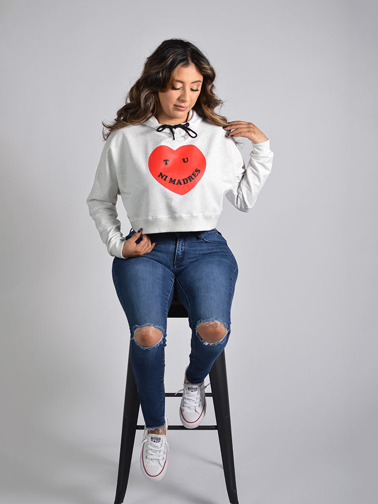 Person wearing a light gray cropped hoodie with a graphic design of a red heart with the text "Tu ni madres" printed on the front