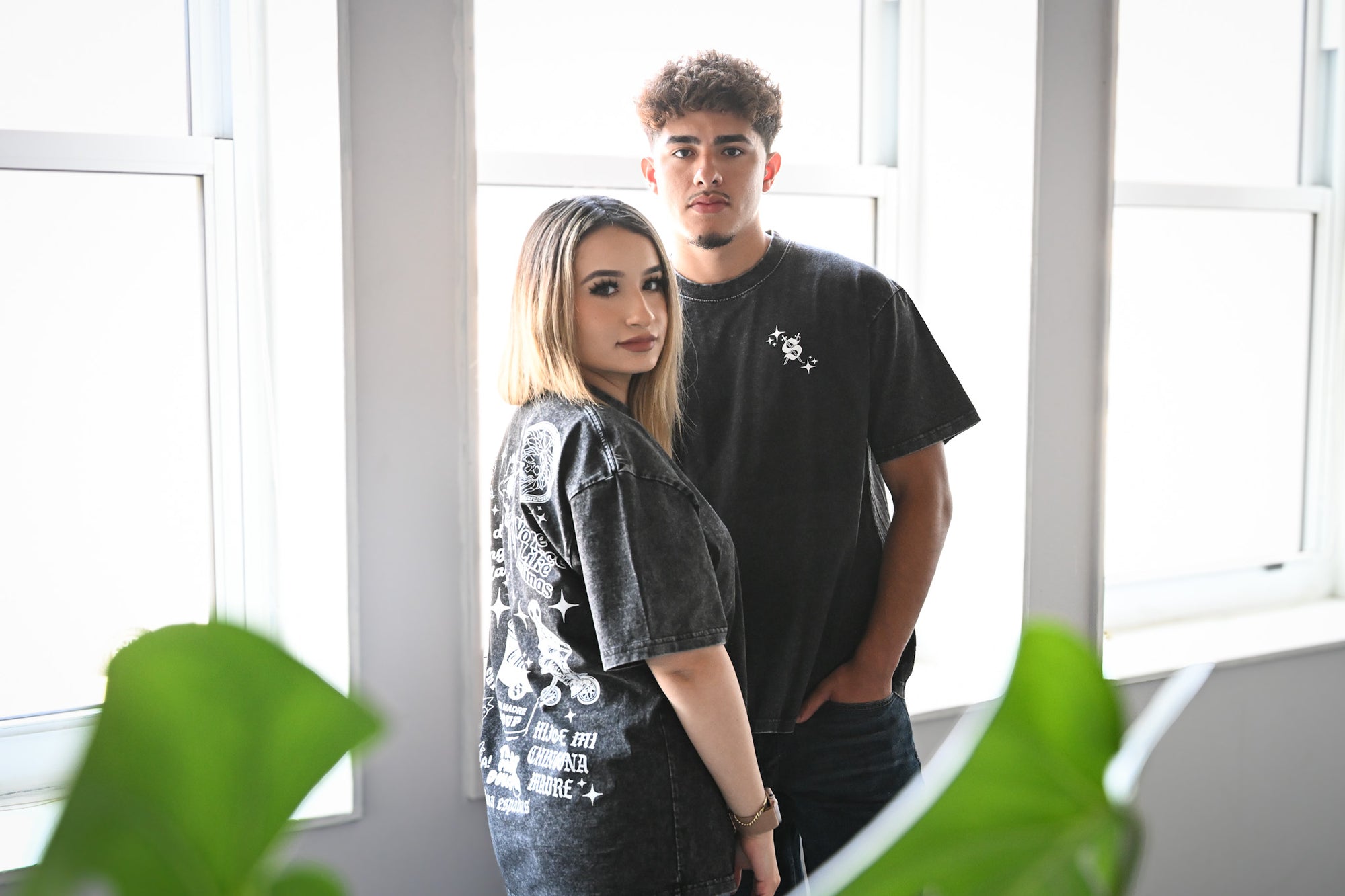 Two people standing in a bright room with large windows and greenery.