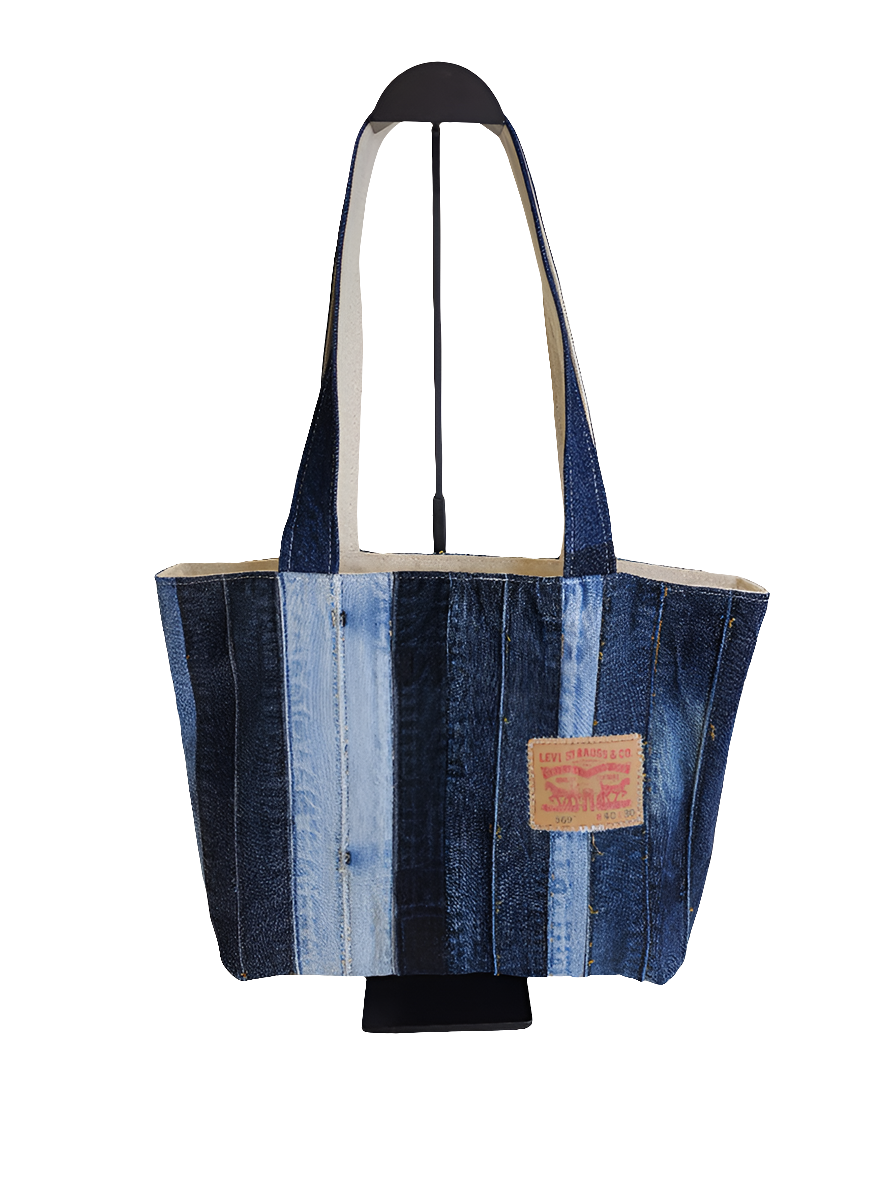 A repurposed blue denim patchwork tote bag with a strap handle, featuring distressed details and a brand tag on the lower side.