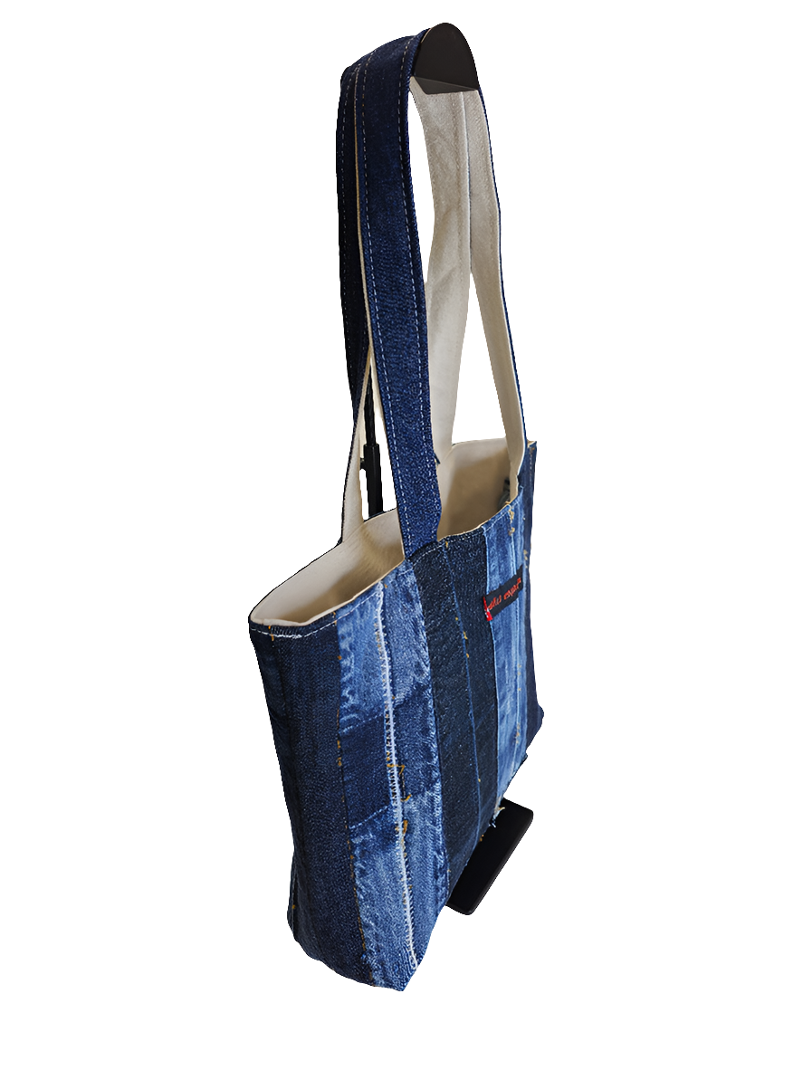 side view of a blue denim patchwork tote bag with a black strap, featuring distressed details and a brand tag on the lower side.