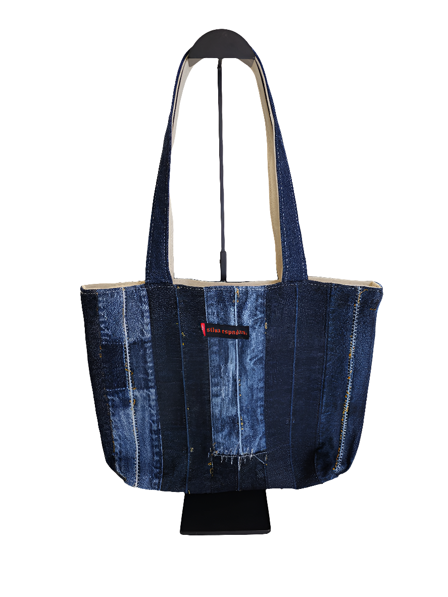 A blue denim patchwork tote bag with a black strap, featuring distressed details and a brand tag on the lower side.