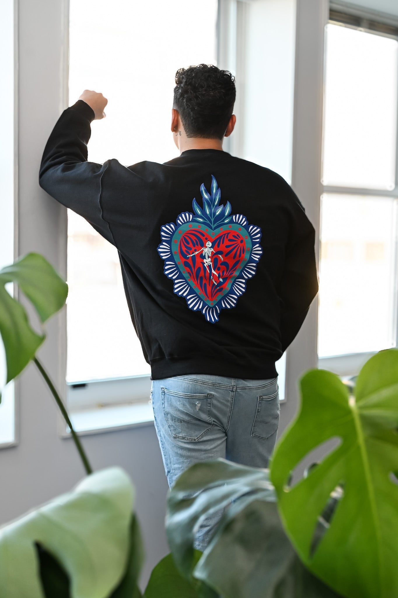 Person wearing a black sweatshirt with a colorful talavera heart and skeleton design, standing indoors with plants in the foreground.