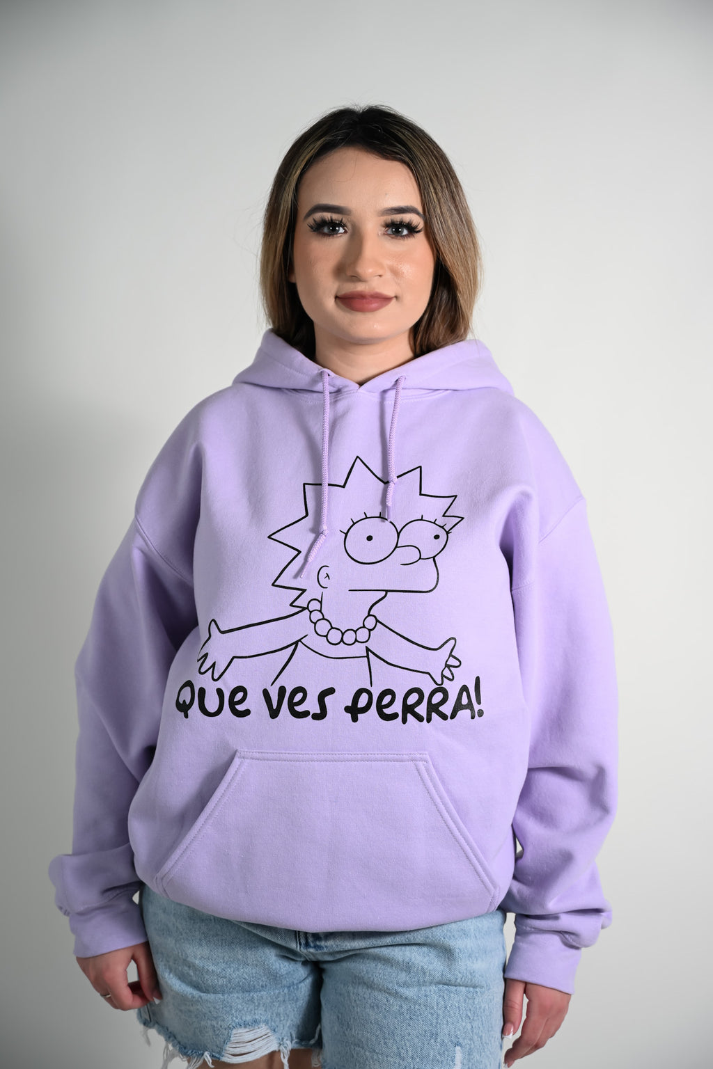 Person wearing a purple hoodie with a cartoon Lisa character and the spanish phrase "Que ves perra" on a plain background