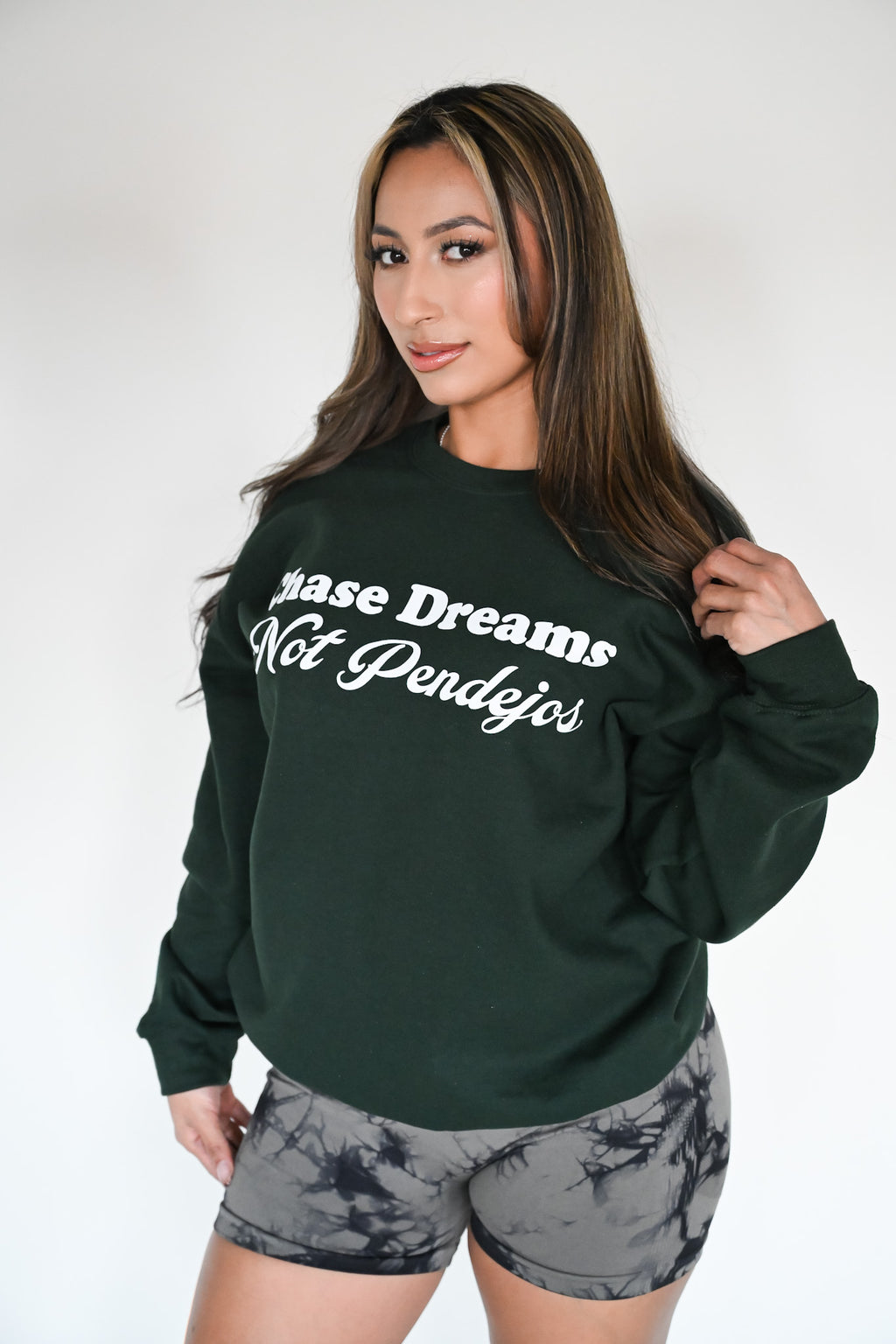 Latina model wearing a green sweater with the phrase " Chase Dreams Not Pendejos" on it