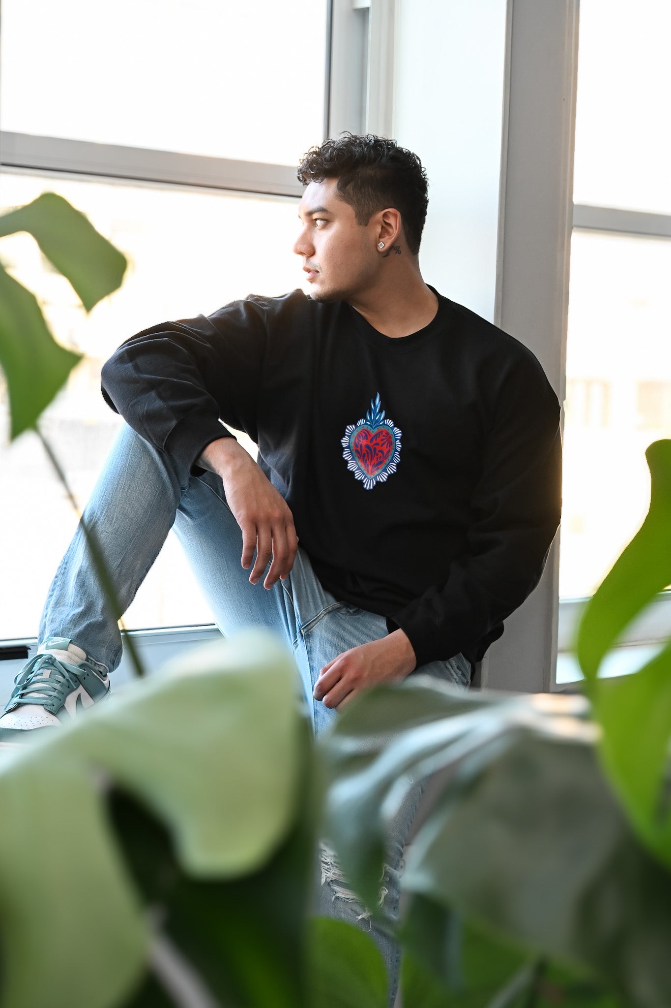 Person wearing a black sweatshirt with a colorful heart design, sitting by a window with greenery.