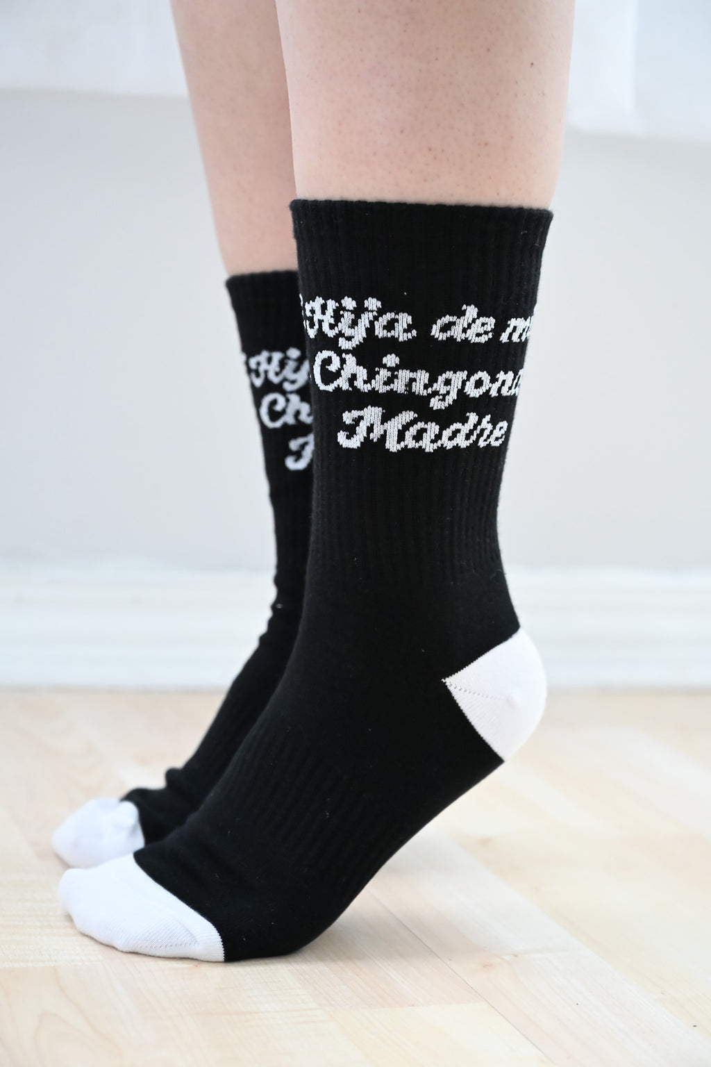 Black Hija de mi chingona madre socks with white text worn by a person on a light wooden floor.