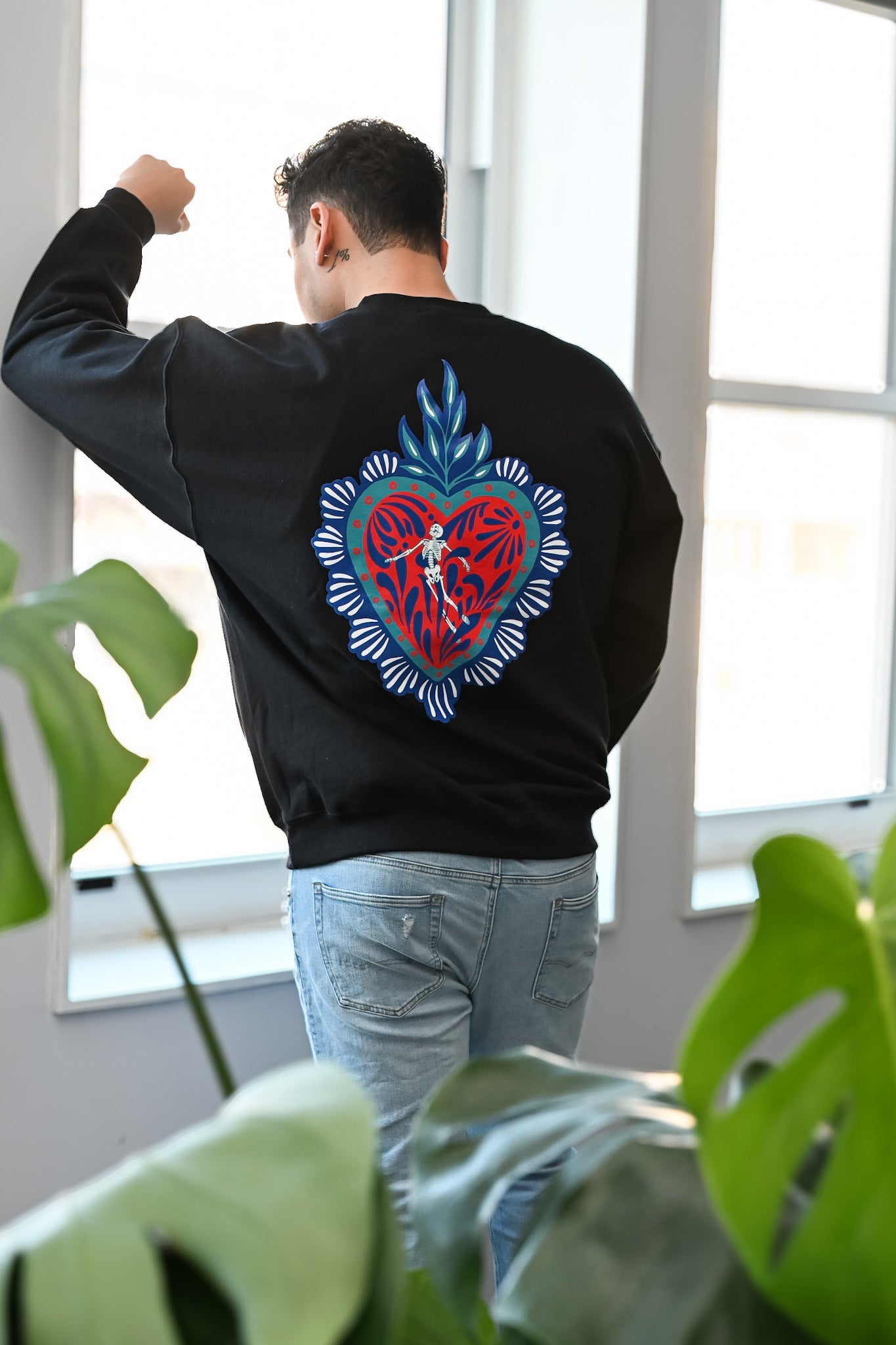 Person wearing a black sweatshirt with a colorful heart design on the back, standing indoors with plants in the foreground.