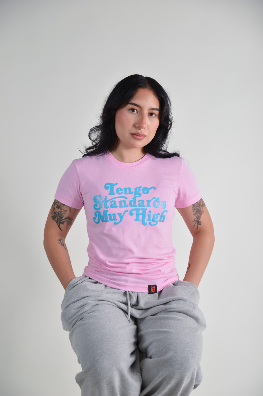 Person wearing a pink t-shirt with blue text "Tengo standards muy high" on a plain background