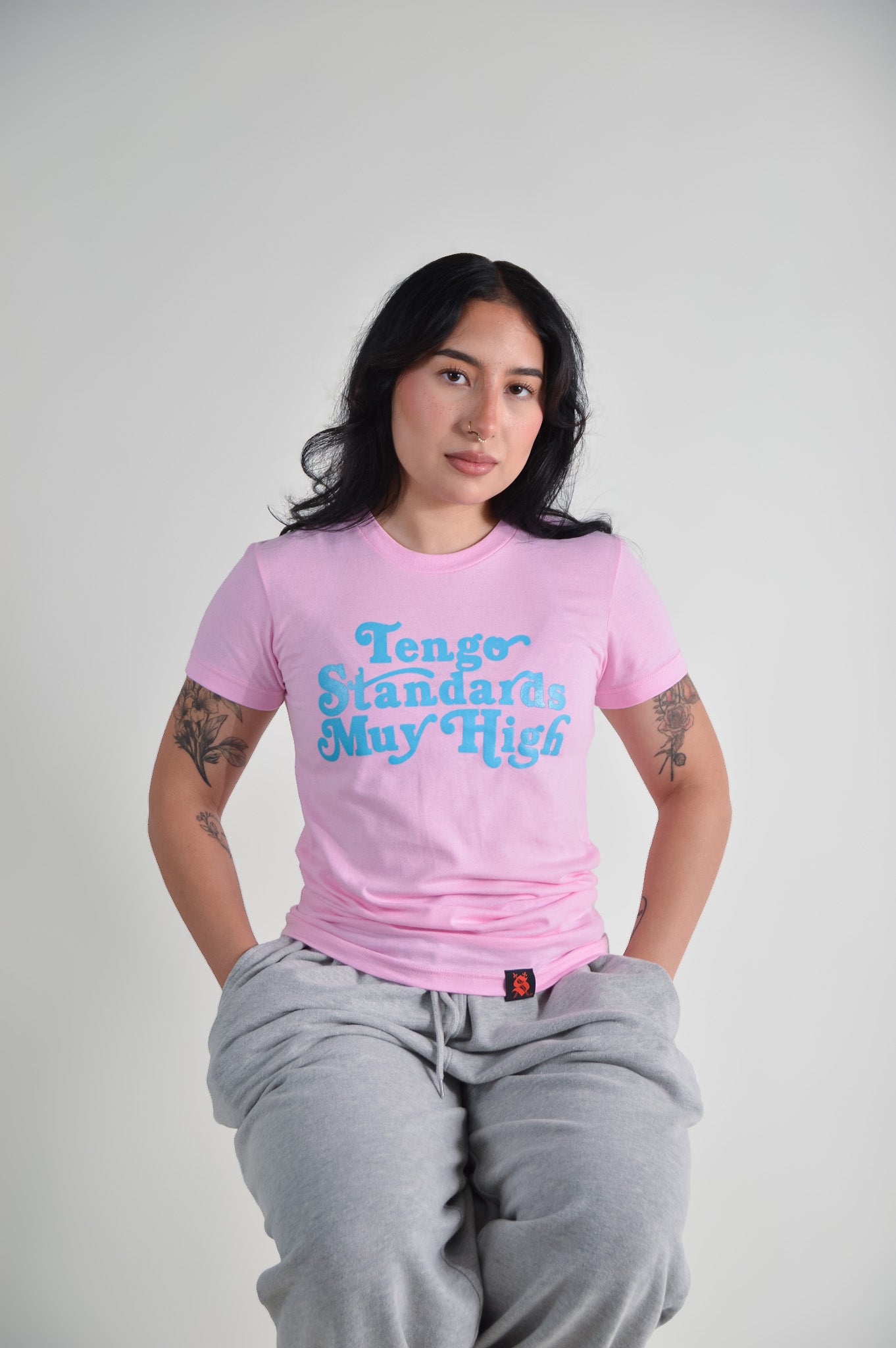 Person wearing a pink t-shirt with blue text "Tengo standards muy high" on a plain background