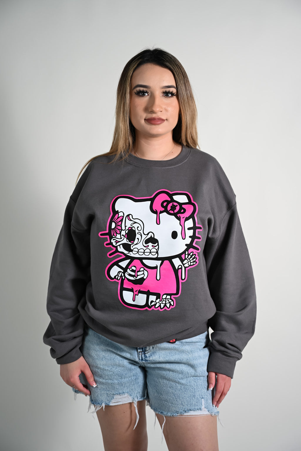 Person wearing a gray sweatshirt with a graphic design of a Hola Kitty day of the dead character in pink and white.