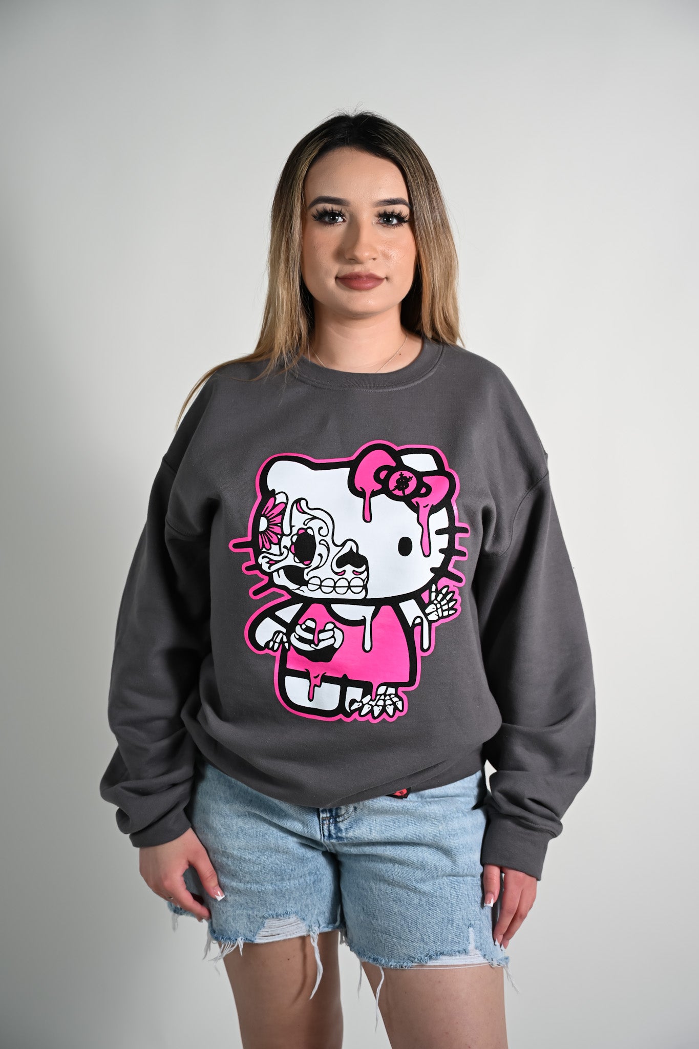 Person wearing a gray sweatshirt with a graphic design of a Hola Kitty day of the dead character in pink and white.