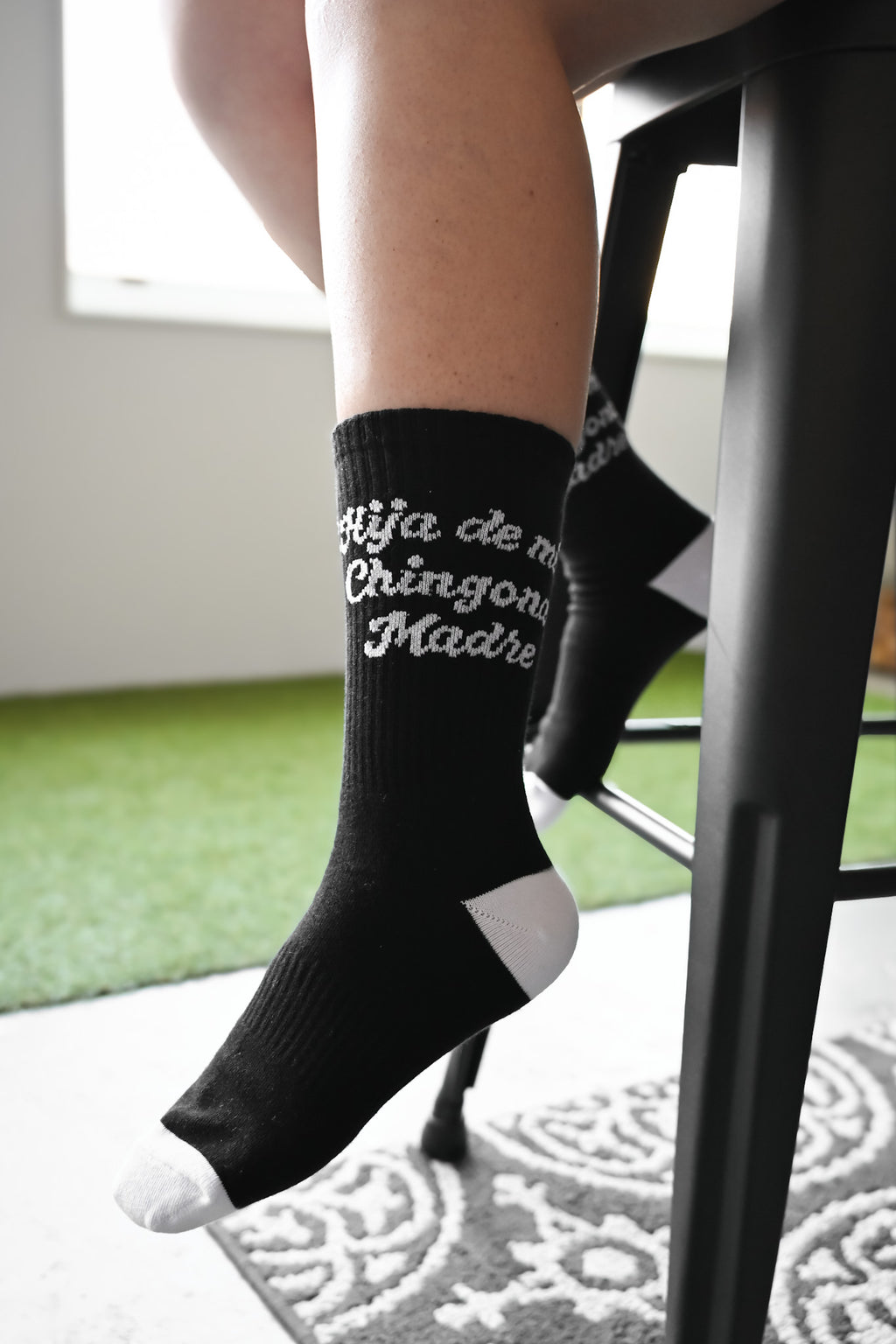 Person wearing black Hija de mi chingona madre socks with white text sitting on a chair.