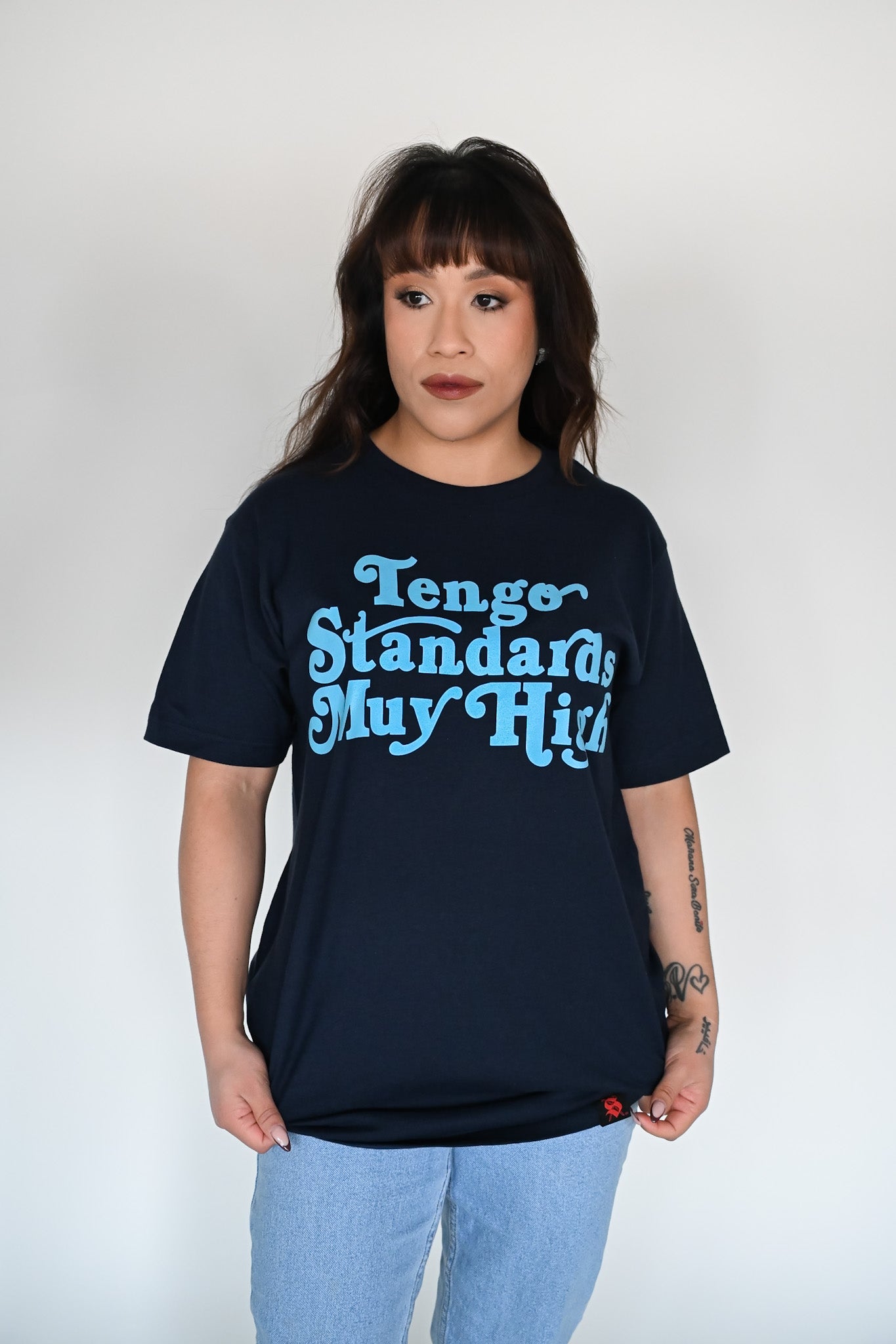 Person wearing a navy blue unisex t-shirt with the phrase "Tengo standards muy high" screen printed on the front