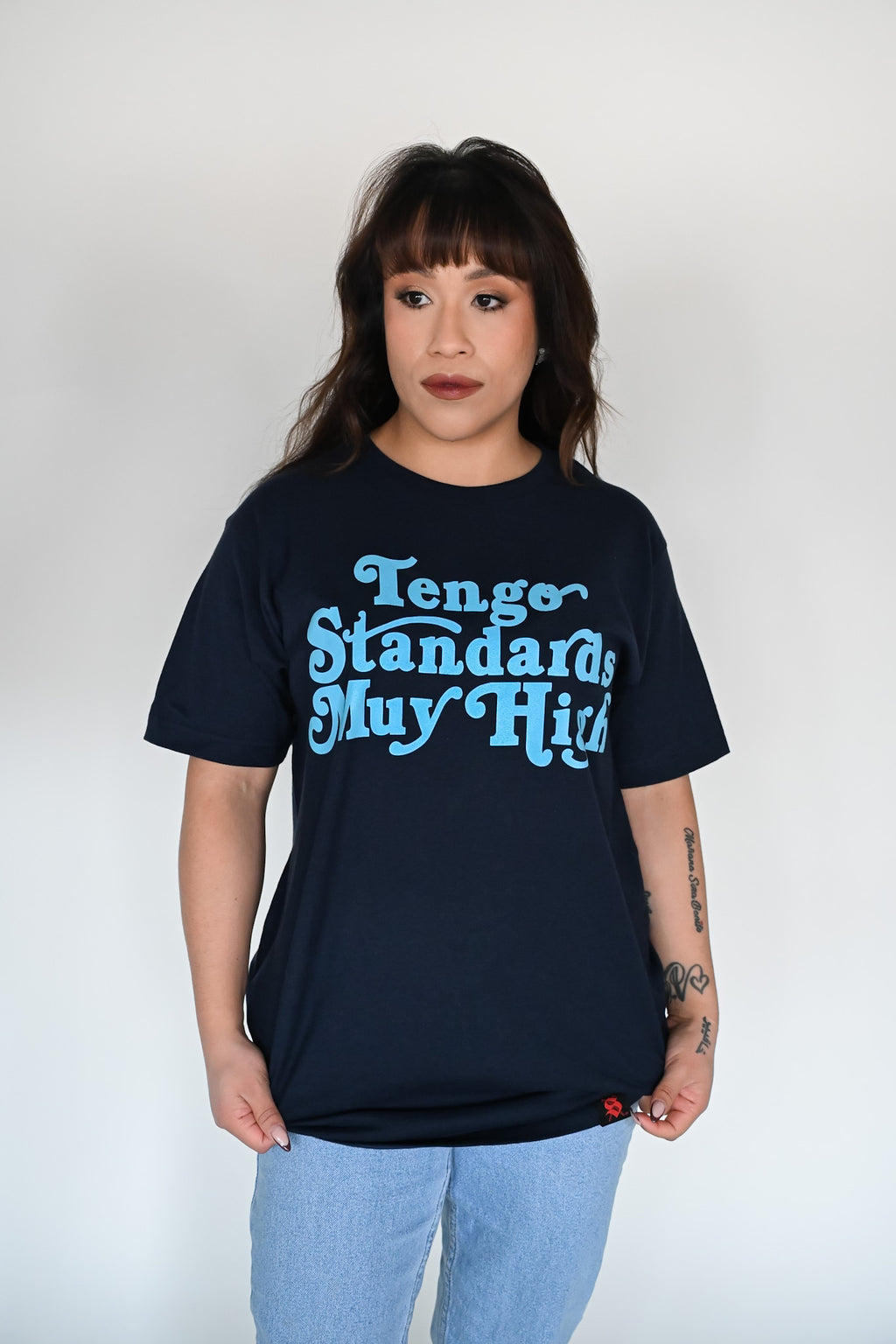 Person wearing a navy blue unisex t-shirt with the phrase "Tengo standards muy high" screen printed on the front