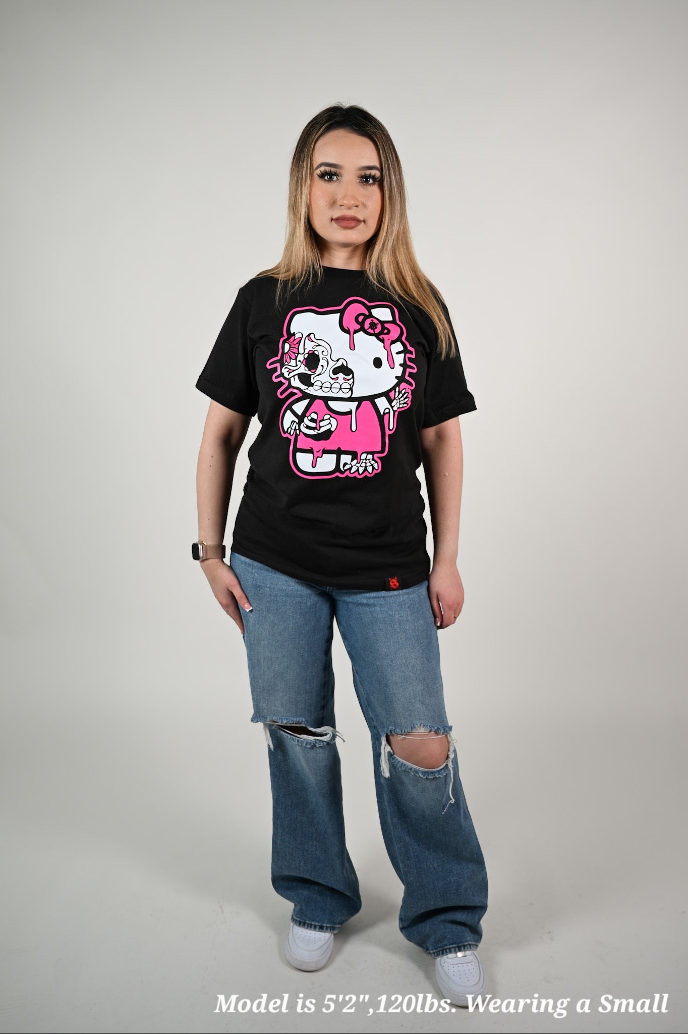 Person wearing a black t-shirt with a hola kitty graphic design and ripped jeans on a gray background