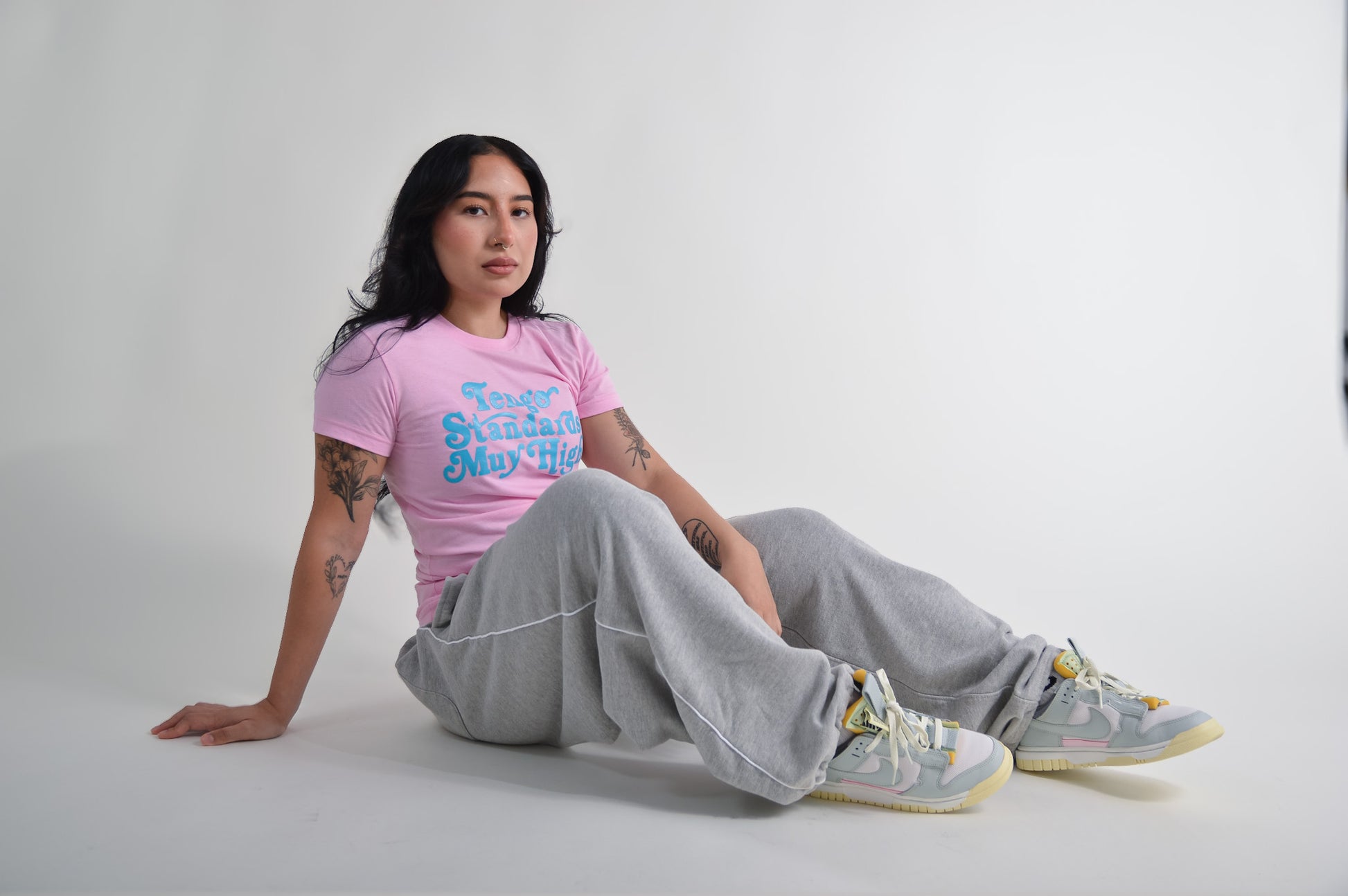 Latina model with tattoos wearing a pink t-shirt with text and gray pants sitting on a white background