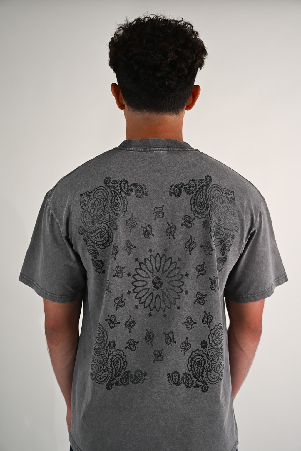 Person wearing a gray t-shirt with black paisley calavera bandana design on the back against a plain background