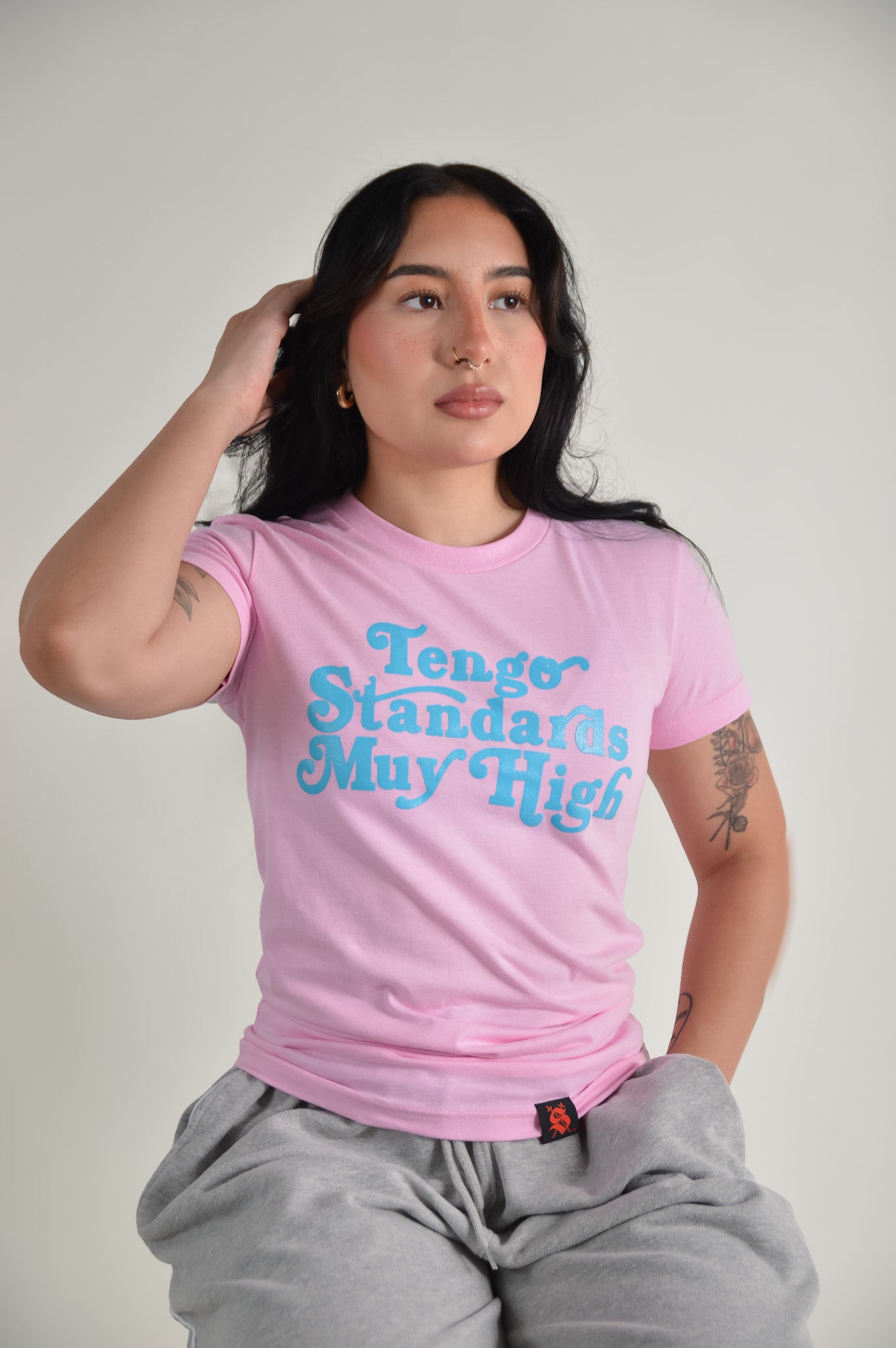 Person wearing a pink t-shirt with blue text "Tengo standards muy high" against a plain background